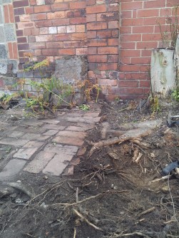 Packing small bricks mark the edge of the brick path, by the tree stump and an iron plate