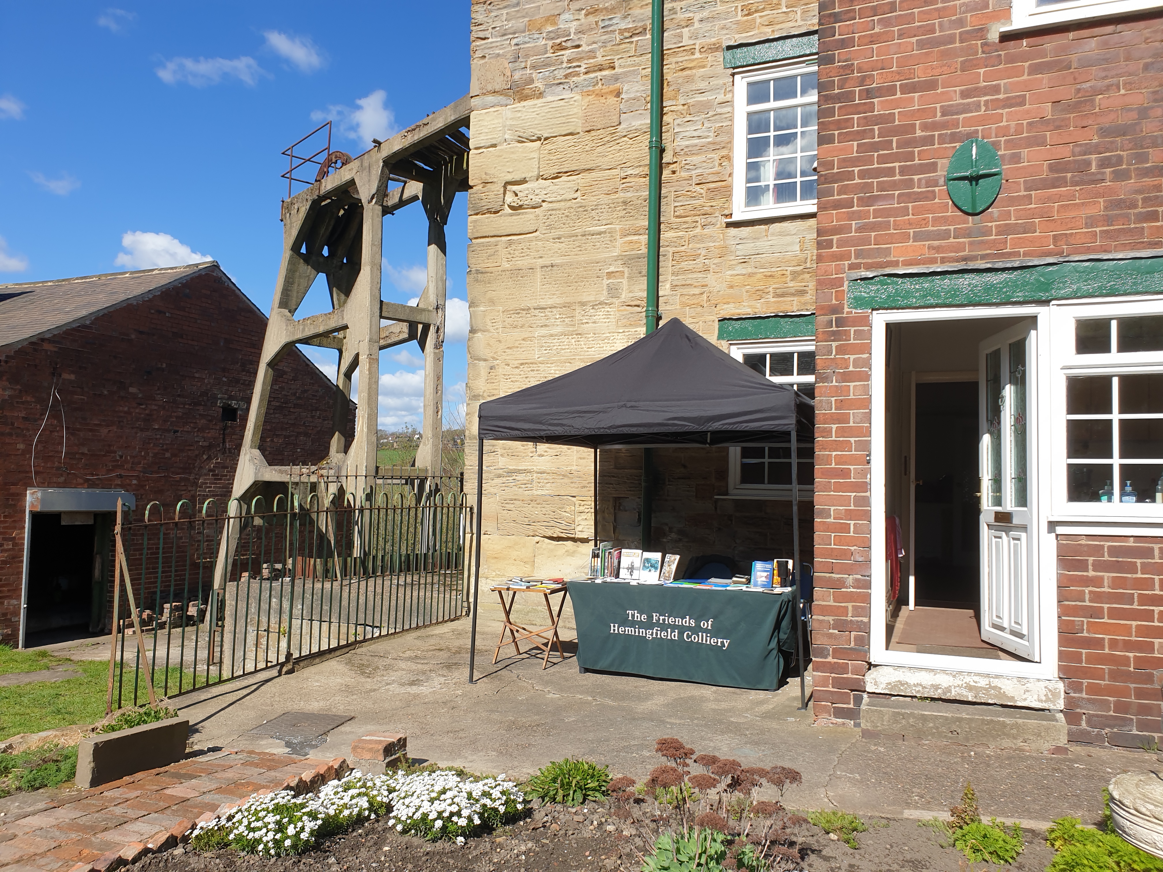 View of a bright sunny day outside Pump House Cottage, with a gazebo and embroidered tablecloth stall set out with books and information for visitors