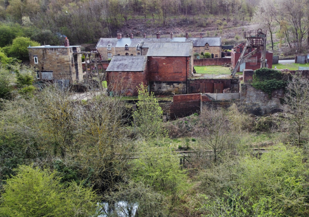 A low-level drone side view of Hemingfield Colliery site, at the top of the photograph a range of buildings fill the shot, the old pumping engine house on the left, the winding engine house in the middle, and roofless surface haulage building to the right. In the foreground the canal basin can be seen through trees, and in the middle across the picture is the line of the railway, currently out of use.