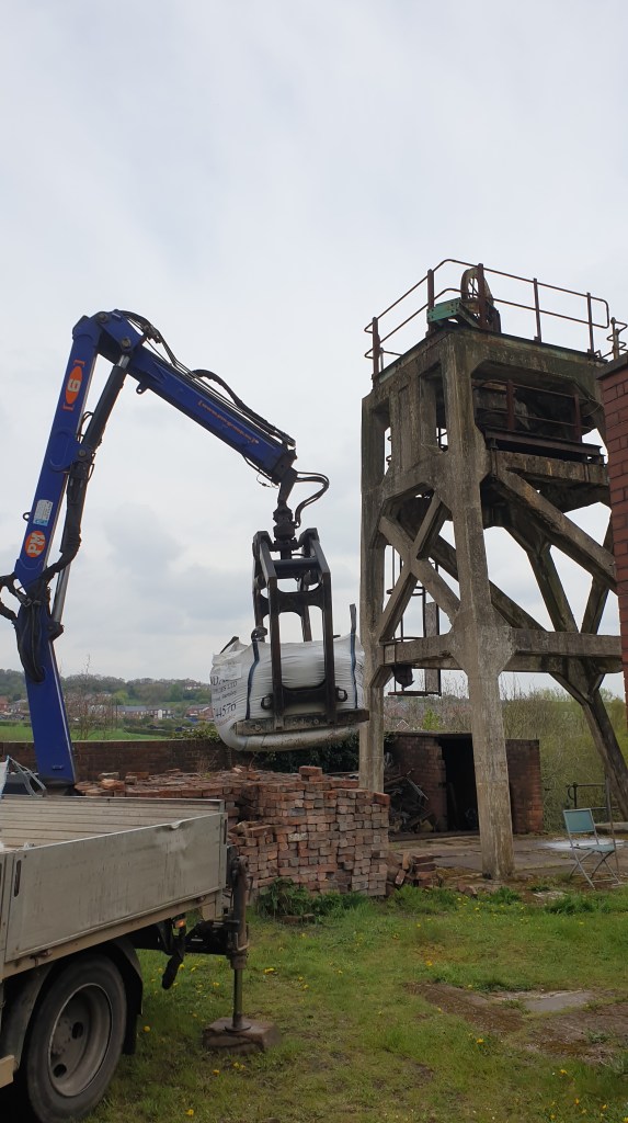 View of a hydraulic lifting arm lowering a builder's merchant sack of sand next to the concrete headgear