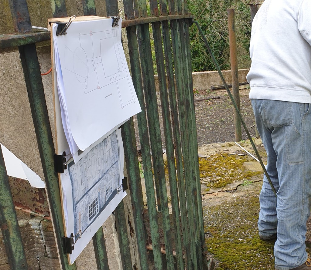 On the left is a clipboard with several sheets of printed paper visible, including a measured drawn plan view of the pumping engine shaft and engine house.  Underneath there is a photographic plan of the masonry of the engine house, to the right can be glimpsed a volunteer in the process of measuring the site.