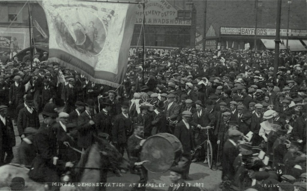 A photograph of a large crowd in the streets of Barnsley watching a public demonstration by the Yorkshire Miners Association. In the foreground a mounted policeman can be seen on horseback, as a march of union men walks through (left-to-right in the image) the scene, led by a marching band. There is a large fabric banner being held high.