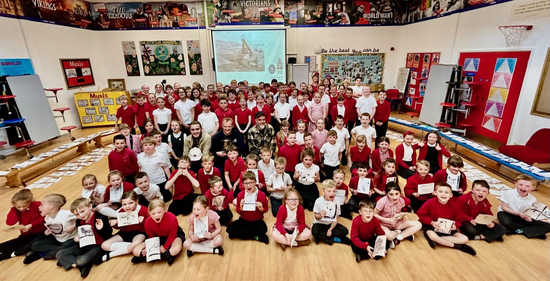 Ellis Primary School, Hemingfield, School assembly jn May 2023 with pupils surrounded by artwork. FOHC Volunteer Mitchell Sutherland, School Governor Mark Farnsworth and Artist Fabric Lenny in the centre.