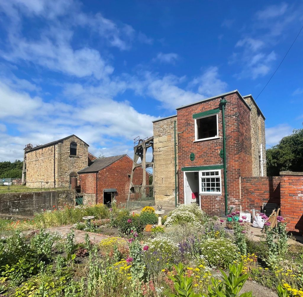 Bright blue sky over Hemingfield Colliery - the winding engine and pumping engine houses in the mid-ground, with the colourful flowers of Pump House Cottage in the foreground.