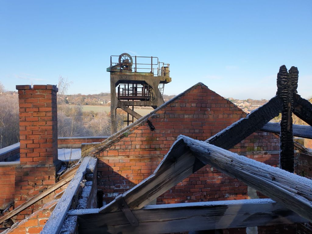 Rooftop view from a roofless burnt out building, looking through burnt timbers and brickwork towards the main concrete headgear at Hemingfield Colliery