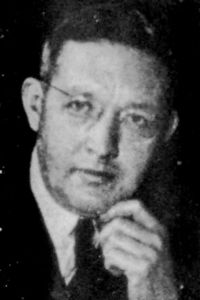 Black and white portrait photograph of Harold Saul (1904-1974) in 1961, wearing glasses, suit jacket and tie with left hand against the left side of his chin.